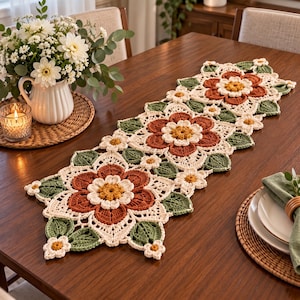 Puede incluir: Un camino de mesa rectangular de ganchillo con un patrón floral repetitivo. El diseño consta de grandes flores de color naranja óxido con centros amarillos, rodeadas de pétalos color crema y hojas de ganchillo verde oscuro.