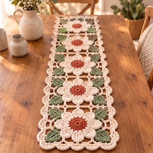 Puede incluir: Un camino de mesa rectangular de ganchillo con un patrón repetitivo de flores color crema con centros color óxido y hojas verde salvia. El camino de mesa tiene un borde festoneado y está construido con motivos florales interconectados.