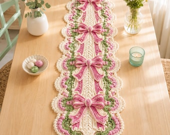 May include: A long, rectangular crochet table runner featuring a cream-colored base with intricate scalloped edges in shades of pink and green. The runner is decorated with four evenly spaced, three-dimensional pink and cream crocheted bows.