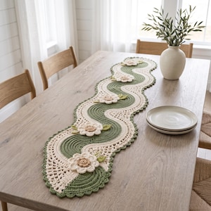Può includere: Un lungo runner da tavola all'uncinetto ondulato con fasce alternate di filato verde salvia e crema. Il runner è decorato con diversi fiori all'uncinetto tridimensionali color crema con centri beige e piccole foglie verdi applicate.
