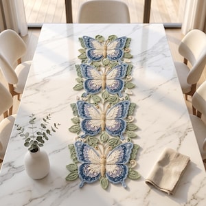 May include: A long, rectangular crochet table runner featuring four large, symmetrical butterfly motifs in shades of blue, cream, and light purple, surrounded by intricate green crocheted leaves and small white floral accents.