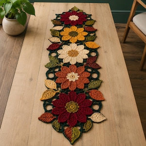 May include: A long, rectangular crochet table runner featuring a series of large, multi-colored floral motifs in shades of deep red, golden yellow, and cream, interspersed with dark green crocheted leaves and intricate openwork circular patterns.