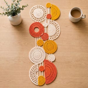 May include: A decorative table runner composed of interconnected crochet circles and semi-circles in cream, orange, and yellow yarn. The geometric design features intricate lace-like patterns and solid stitches, arranged in a vertical, elongated composition.