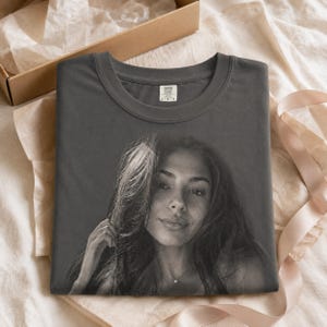 May include: A charcoal gray crew neck t-shirt featuring a large, monochromatic photographic print of a person with long hair on the front. The shirt has a visible interior neck label that reads Comfort Colors and the size indicator L.