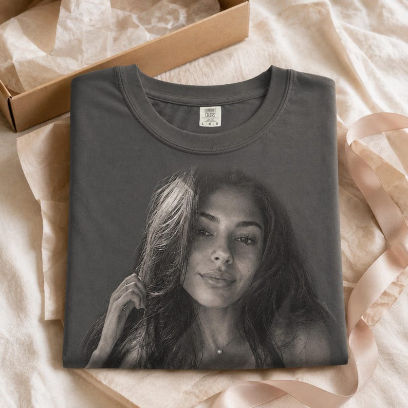 May include: A charcoal gray crew neck t-shirt featuring a large, monochromatic photographic print of a person with long hair on the front. The shirt has a visible interior neck label that reads Comfort Colors and the size indicator L.