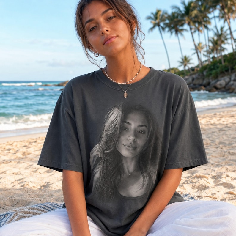 May include: A charcoal gray short-sleeve t-shirt featuring a large, centered black and white photographic print of a person's face. The shirt has a crew neckline and a relaxed, boxy fit, constructed from a solid-colored cotton fabric.