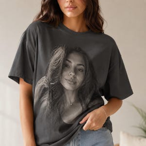 May include: A charcoal gray short-sleeve t-shirt featuring a large, centered black and white photographic print of a person with long hair. The shirt has a crew neckline, a relaxed fit, and a smooth, solid-colored fabric construction throughout.