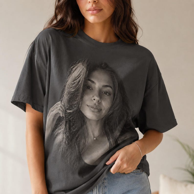 May include: A charcoal gray short-sleeve t-shirt featuring a large, centered black and white photographic print of a person with long hair. The shirt has a crew neckline, a relaxed fit, and a smooth, solid-colored fabric construction throughout.