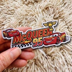 Può includere: Una toppa ricamata raffigurante il personaggio dell'auto da corsa rossa Lightning McQueen a sinistra e a destra, con il testo McQueen 95 in rosso e giallo, un'icona di trofeo dorato in alto e un motivo a scacchi bianco e nero da corsa.