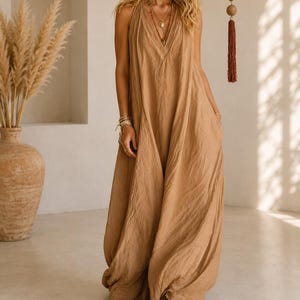 May include: A floor-length, sleeveless tan dress with a deep V-neckline and a loose, flowing silhouette. The garment features a textured, slightly wrinkled fabric finish, wide shoulder straps, and side pockets, extending down to the floor.