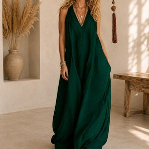 May include: A floor-length, dark green sleeveless dress with a deep V-neckline and a loose, flowing silhouette. The garment features a textured fabric finish, a halter-style bodice, and a long, wide skirt that reaches the floor, paired with brown shoes.