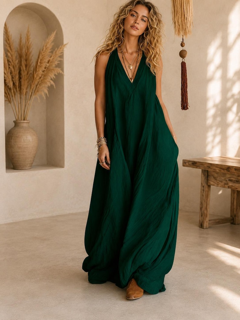 May include: A floor-length, dark green sleeveless dress with a deep V-neckline and a loose, flowing silhouette. The garment features a textured fabric finish, a halter-style bodice, and a long, wide skirt that reaches the floor, paired with brown shoes.