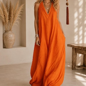 May include: A vibrant orange sleeveless maxi dress featuring a deep V-neckline and a loose, flowing silhouette that reaches the floor. The garment is made of a textured fabric and includes side pockets, paired with brown pointed-toe ankle boots.