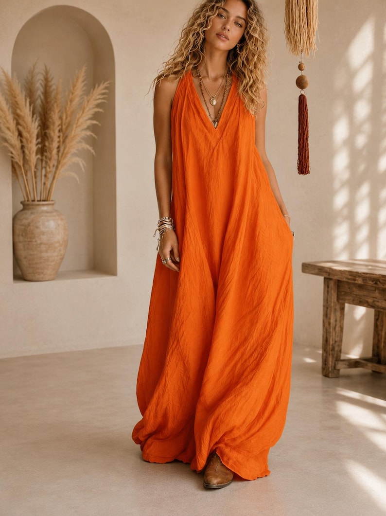 May include: A vibrant orange sleeveless maxi dress featuring a deep V-neckline and a loose, flowing silhouette that reaches the floor. The garment is made of a textured fabric and includes side pockets, paired with brown pointed-toe ankle boots.