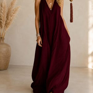 May include: A floor-length, deep burgundy sleeveless dress featuring a deep V-neckline and a loose, flowing silhouette. The garment is made of a textured fabric and includes side pockets, falling to the floor with a relaxed, draped fit.