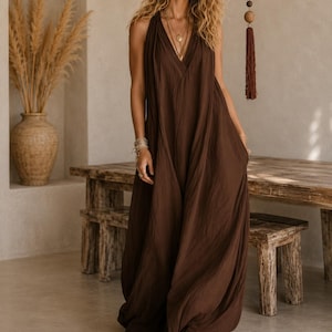 May include: A floor-length, sleeveless dark brown dress with a deep V-neckline and a loose, flowing silhouette. The garment features a gathered bodice, wide shoulder straps, and a long, voluminous skirt that reaches the floor, made of a solid-colored fabric.