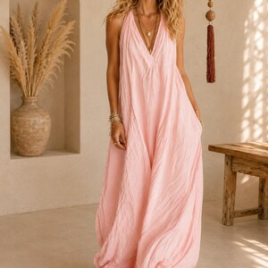 May include: A floor-length, light pink sleeveless dress with a deep V-neckline and a textured, crinkled fabric finish. The garment features a loose, flowing silhouette that drapes to the floor, with wide straps and a simple, unadorned design.