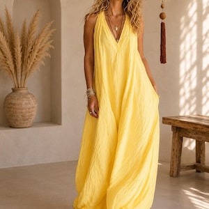 May include: A bright yellow sleeveless maxi dress featuring a deep V-neckline, a loose and flowing silhouette, and a textured fabric finish. The garment falls to the floor and includes layered necklaces and stacked bracelets as accessories.