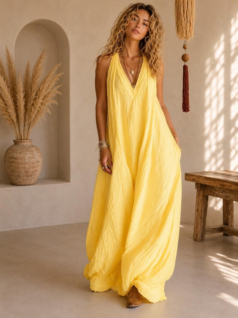 May include: A bright yellow sleeveless maxi dress featuring a deep V-neckline, a loose and flowing silhouette, and a textured fabric finish. The garment falls to the floor and includes layered necklaces and stacked bracelets as accessories.