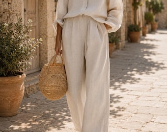 May include: A beige long-sleeved button-down shirt and matching wide-leg trousers made of a textured fabric. The outfit is paired with a round woven straw handbag and simple open-toe sandals, creating a coordinated two-piece ensemble.