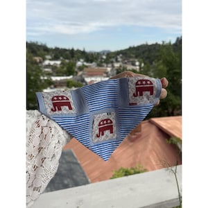 May include: A triangular pet bandana featuring a patchwork design of blue and white horizontal stripes alternating with light gray squares. Each gray square displays a dark red elephant graphic with three white stars above the elephant's back.