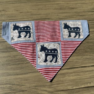 Peut inclure: Un bandana triangulaire pour animal de compagnie avec un motif patchwork composé de rayures horizontales rouges et blanches alternant avec des carrés bleu clair. Chaque carré bleu présente une silhouette d'âne bleu foncé avec quatre étoiles blanches.