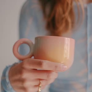Puede incluir: Una taza de cerámica con un cuerpo liso y redondeado que presenta un degradado de color desde un rosa suave en la parte superior hasta un amarillo cálido en la base. La taza incluye un asa circular distintiva que combina con el tono rosa del borde.