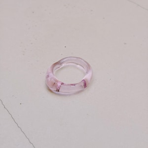 Puede incluir: Un anillo de color rosa claro translúcido con una forma orgánica suave, redondeada y ligeramente irregular. La banda está hecha de un material transparente que permite el paso de la luz, creando reflejos sutiles y profundidad.