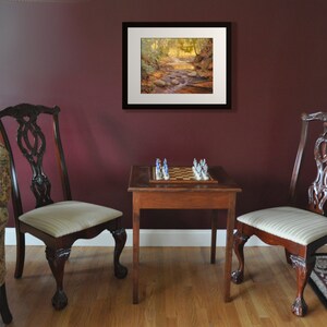 May include: A framed print of a forest scene with a stream running through it. The print is hanging on a maroon wall above a chessboard on a wooden table. Two chairs are positioned on either side of the table.