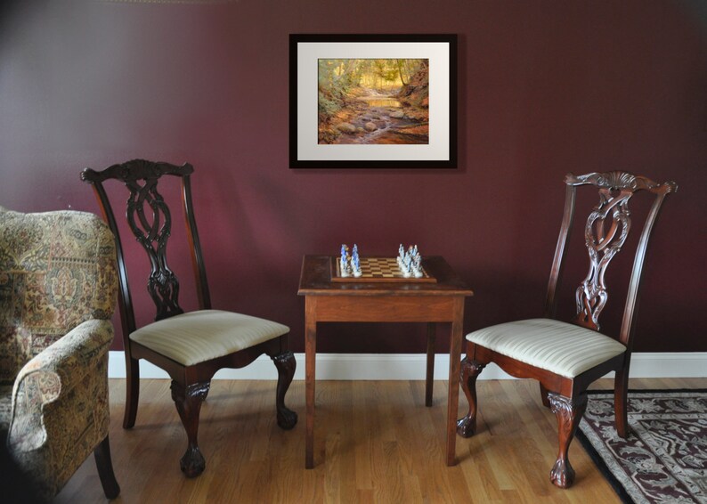 May include: A framed print of a forest scene with a stream running through it. The print is hanging on a maroon wall above a chessboard on a wooden table. Two chairs are positioned on either side of the table.