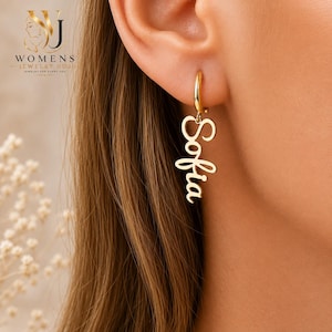 May include: A gold-toned hoop earring featuring a dangling nameplate pendant with the name Sofia written in a cursive script font. The earring is suspended from an earlobe, showcasing a polished metallic finish and a vertical orientation.