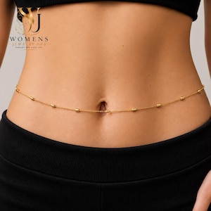 May include: A delicate gold-toned waist chain featuring a thin metal link chain interspersed with small, cylindrical gold beads. The jewelry rests horizontally across the midsection, positioned just above the navel area against the skin.