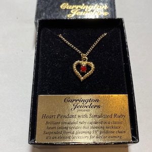 May include: A gold-tone heart pendant necklace featuring a small red simulated ruby at its center. The pendant is suspended from a delicate chain and presented in a black box with a gold label reading Carrington Jewelers Heart Pendant with Simulated Ruby.