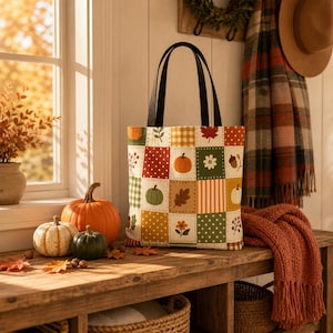 Op de afbeelding: Een draagtas met een patchworkontwerp van herfstachtige vierkanten. Het patroon bevat oranje, groene en witte pompoenen, bruine eikenbladeren, rode esdoornbladeren, witte bloemen en diverse stoffen met stippen en strepen.