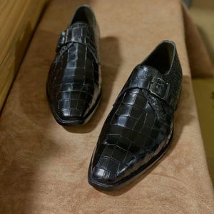 May include: A pair of black monk strap shoes featuring a glossy finish with a prominent embossed crocodile skin texture. Each shoe is designed with a single metallic buckle closure across the top and a pointed toe silhouette with a low heel.