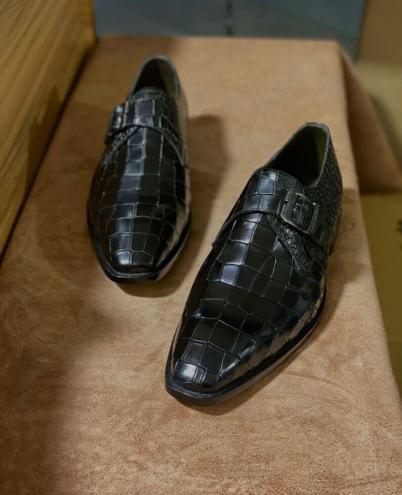 May include: A pair of black monk strap shoes featuring a glossy finish with a prominent embossed crocodile skin texture. Each shoe is designed with a single metallic buckle closure across the top and a pointed toe silhouette with a low heel.