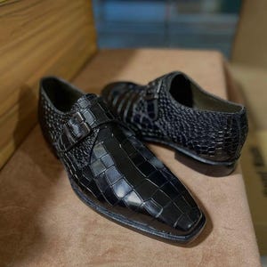 May include: A pair of black monk strap shoes featuring a glossy crocodile-embossed texture. Each shoe has a single silver-toned metal buckle closure on the side, a square toe design, and a low heel, showcasing a polished finish throughout.