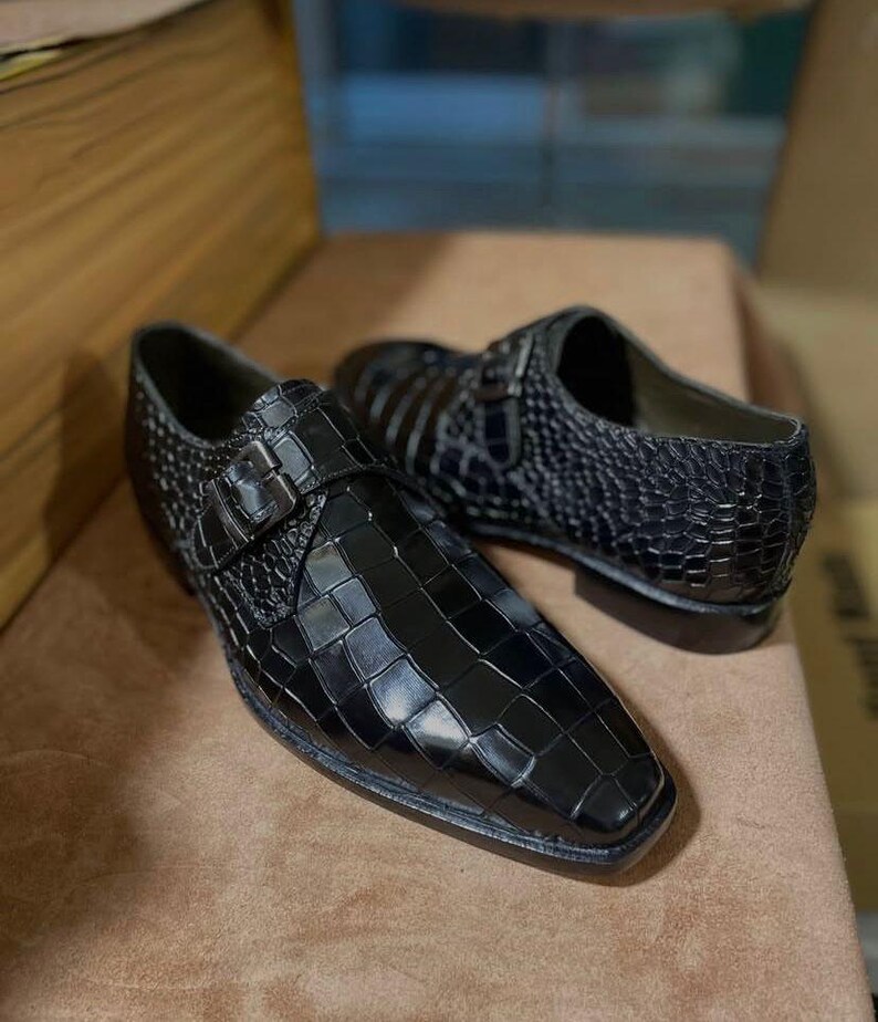 May include: A pair of black monk strap shoes featuring a glossy crocodile-embossed texture. Each shoe has a single silver-toned metal buckle closure on the side, a square toe design, and a low heel, showcasing a polished finish throughout.