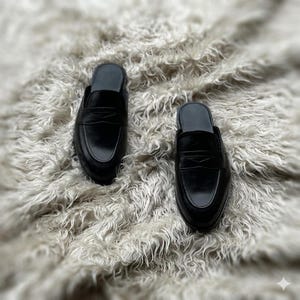 May include: A pair of black leather slip-on loafers featuring a classic penny loafer strap across the vamp, a rounded toe, and an open-back design. The shoes have a smooth, polished finish and a low-profile construction with a simple, clean silhouette.