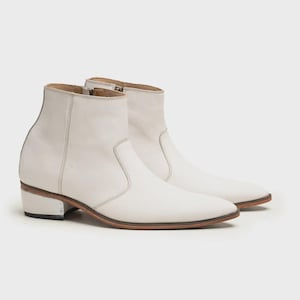 May include: A pair of off-white leather ankle boots featuring a pointed toe, a side zipper closure, and a low stacked heel with a dark brown sole edge. The boots have a smooth finish and a structured silhouette with visible stitching along the panels.