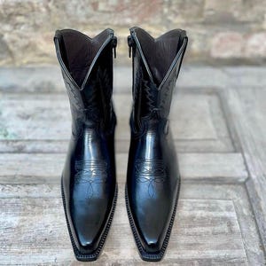 May include: A pair of black leather cowboy boots featuring a pointed toe, a side zipper closure, and decorative tonal stitching on the shaft and vamp. The boots have a structured silhouette with a V-shaped topline and a smooth, polished finish.