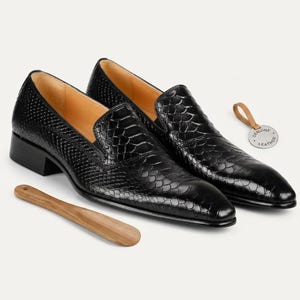 May include: A pair of black slip-on loafers with a textured snakeskin pattern, featuring a tan interior lining and a low block heel. A small round tag labeled Genuine Leather is attached by a tan loop, and a wooden shoehorn lies in the foreground.