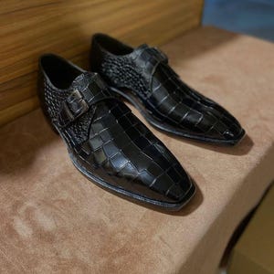 May include: A pair of black dress shoes with a glossy crocodile-embossed texture. Each shoe features a pointed toe, a single side strap with a metallic buckle closure, and a low heel. The design includes a structured silhouette with a polished finish.
