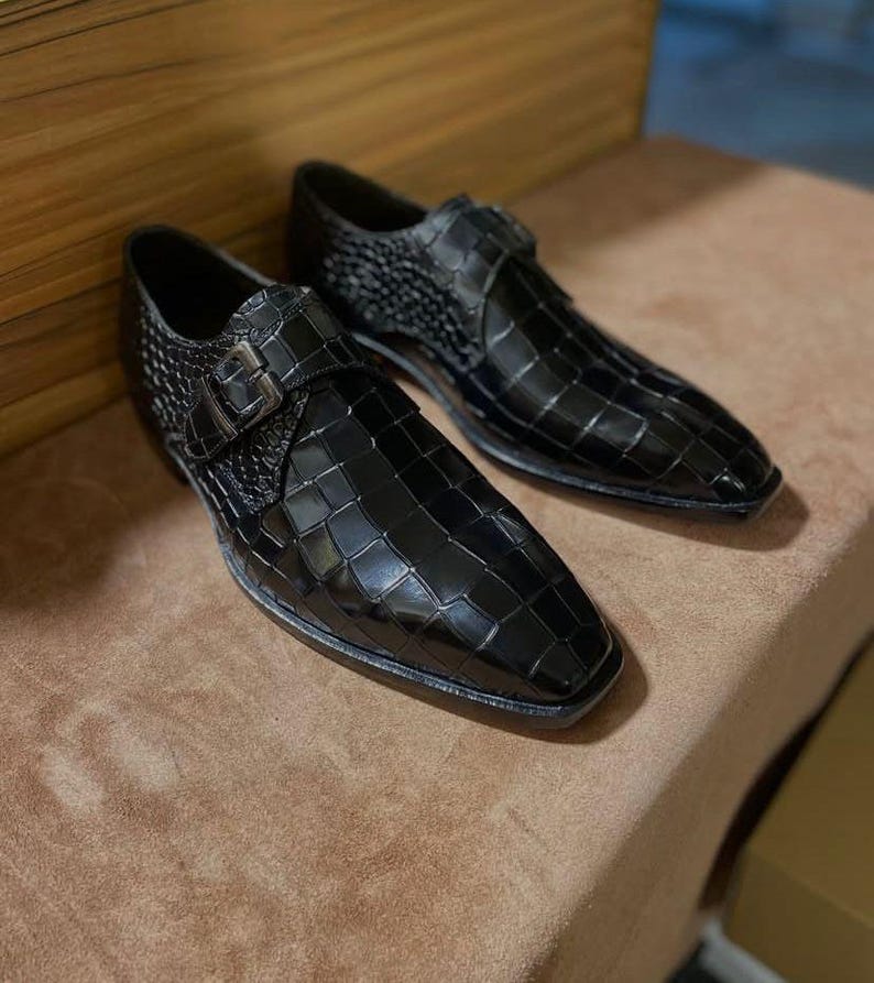 May include: A pair of black dress shoes with a glossy crocodile-embossed texture. Each shoe features a pointed toe, a single side strap with a metallic buckle closure, and a low heel. The design includes a structured silhouette with a polished finish.