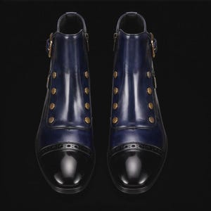 May include: A pair of navy blue ankle boots featuring a glossy black cap toe with brogue detailing. The boots have a side buckle strap closure and a vertical row of four decorative gold-toned buttons on each side of the front shaft.