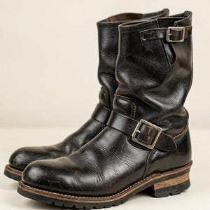 May include: A pair of black leather engineer boots featuring a rounded toe, a mid-calf shaft, and two adjustable straps with silver-toned metal buckles. The boots have a stacked wooden heel and a thick, lugged rubber outsole for traction.