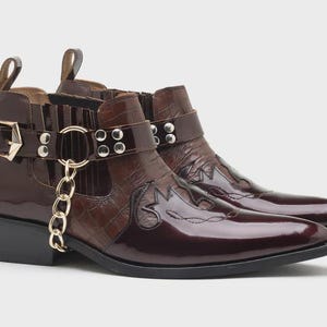 May include: A pair of dark brown ankle boots featuring a glossy finish, a crocodile-embossed texture on the side panels, and a decorative strap with silver-tone metal studs, a buckle, and a dangling gold-tone chain link accent on the side.