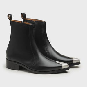 May include: A pair of black ankle boots featuring a smooth finish, a low block heel, a side zipper closure, and a metallic silver-toned cap toe detail. The boots have a pull tab at the back of the collar and a structured, pointed toe silhouette.