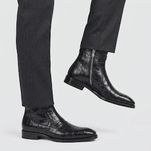Könnte beinhalten: Ein Paar schwarze Stiefeletten mit einer strukturierten Oberfläche in Krokodilleder-Optik. Die Stiefel haben eine schlanke Silhouette mit einem niedrigen Blockabsatz, einer spitzen Zehenpartie und einem vertikalen silberfarbenen Reißverschluss.
