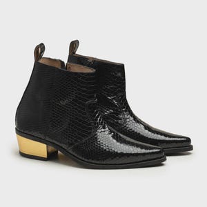 Könnte beinhalten: Ein Paar schwarze Stiefeletten mit texturiertem Schlangenledermuster, spitzer Zehenpartie und einem metallisch goldenen Blockabsatz. Jeder Stiefel verfügt über eine Zuglasche am hinteren Schaft und einen seitlichen Reißverschluss mit glänzendem Finish.